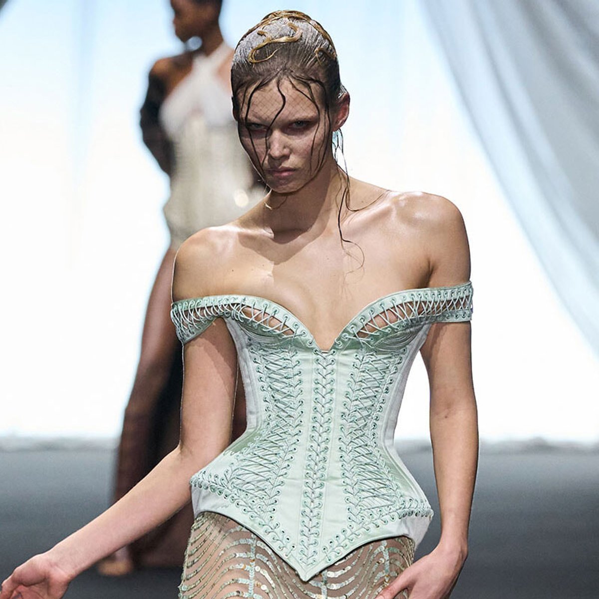 Model walking a fashion runway in a pale mint, off-shoulder corset with intricate lattice lacing and sheer lower panel, wet hair pulled back.