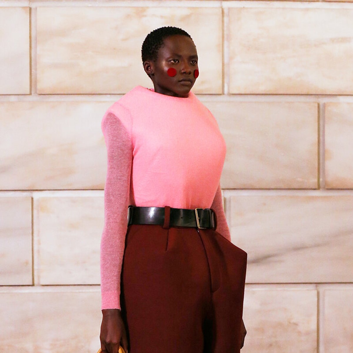 Person in a bright pink knit top and high-waisted burgundy pants with a black belt, posed against a light stone wall (red circular cheek makeup visible).