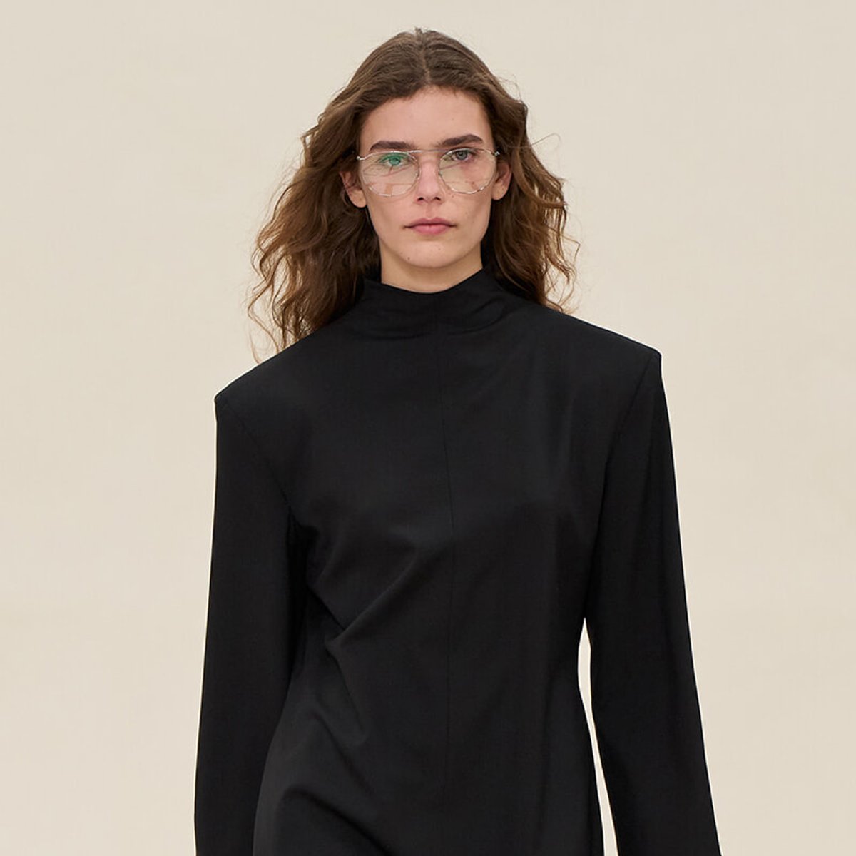 Woman wearing a black high-neck dress and clear glasses against a beige background, posing for a fashion shot.