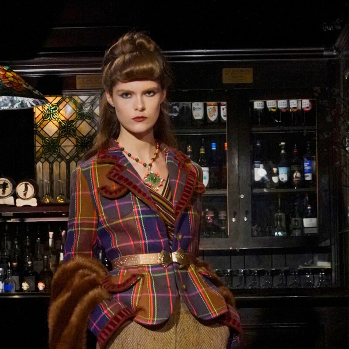 Model wearing a multicolored plaid coat with fur cuffs stands in a bar setting with bottles in the background.