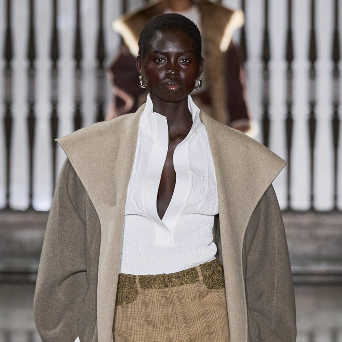 Model walks a fashion runway in a white blouse with a large collar, beige coat draped over shoulders, and tan plaid trousers with a gold belt.