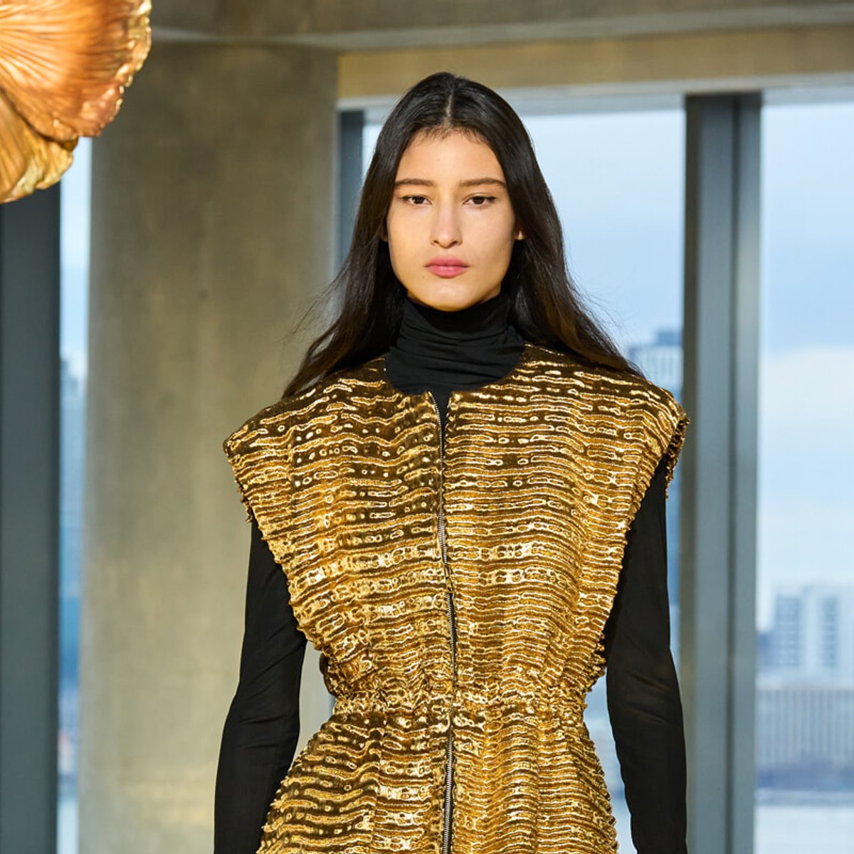 Model wearing a metallic gold textured vest over a black turtleneck, standing in a modern indoor setting during a fashion show.