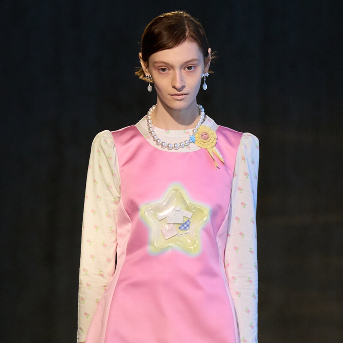 Model on a runway wearing a pink satin dress over a white floral long-sleeve top, accessorized with a pearl necklace and drop earrings, and a yellow rosette badge on the shoulder.