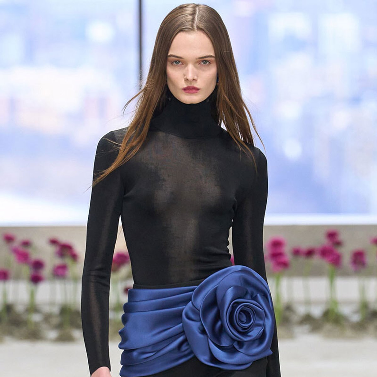 Model wearing a sheer black turtleneck with a bold blue satin sash shaped into a rose at the waist, on a runway backdrop.