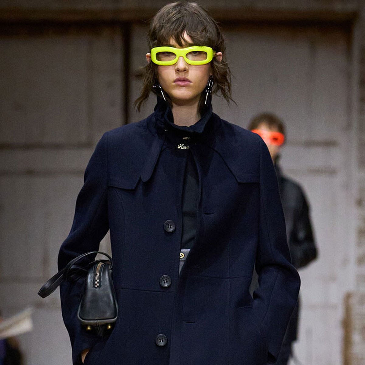 Model on a runway wearing a navy oversized coat, neon yellow sunglasses, and carrying a black handbag.