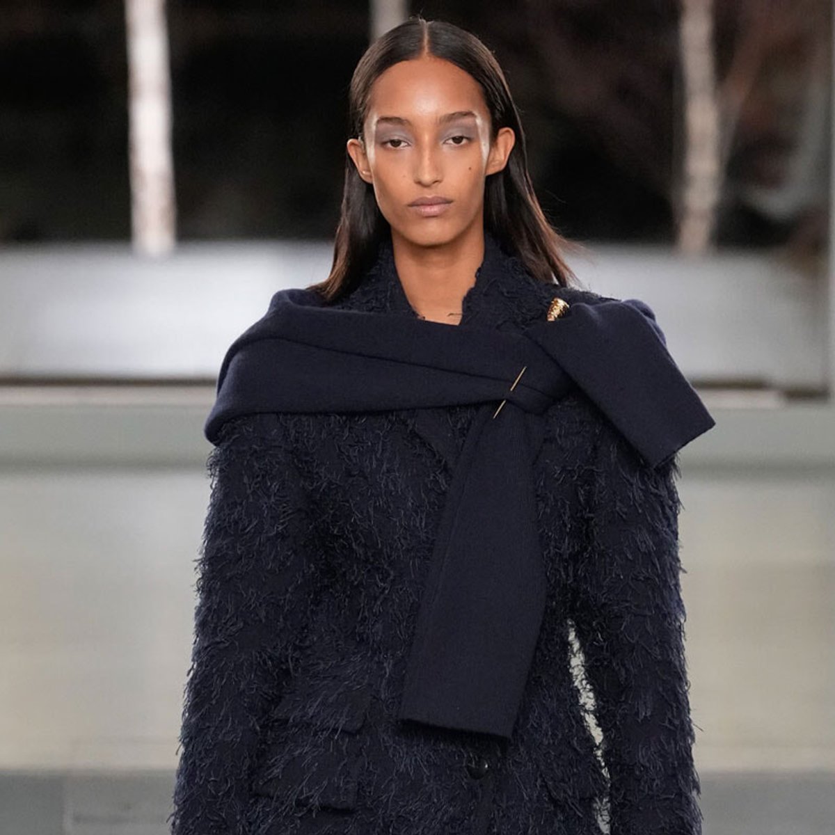 Female model on a runway wearing a dark navy textured coat with a long navy scarf wrapped across the shoulders and chest.