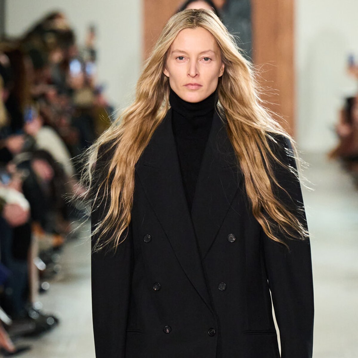 Model walking a runway in a black double-breasted coat with a turtleneck, long blonde hair flowing.