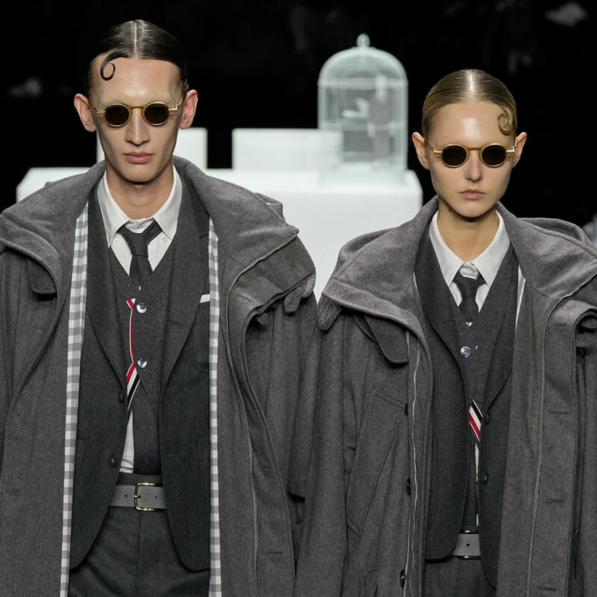 Two models on a fashion runway wearing oversized gray coats over suits, with round sunglasses and slicked-back hair.
