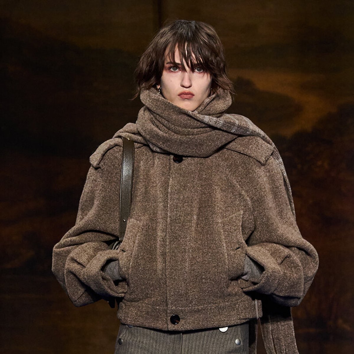 Model wearing an oversized brown textured coat with a chunky scarf and shoulder bag, against a dark background.