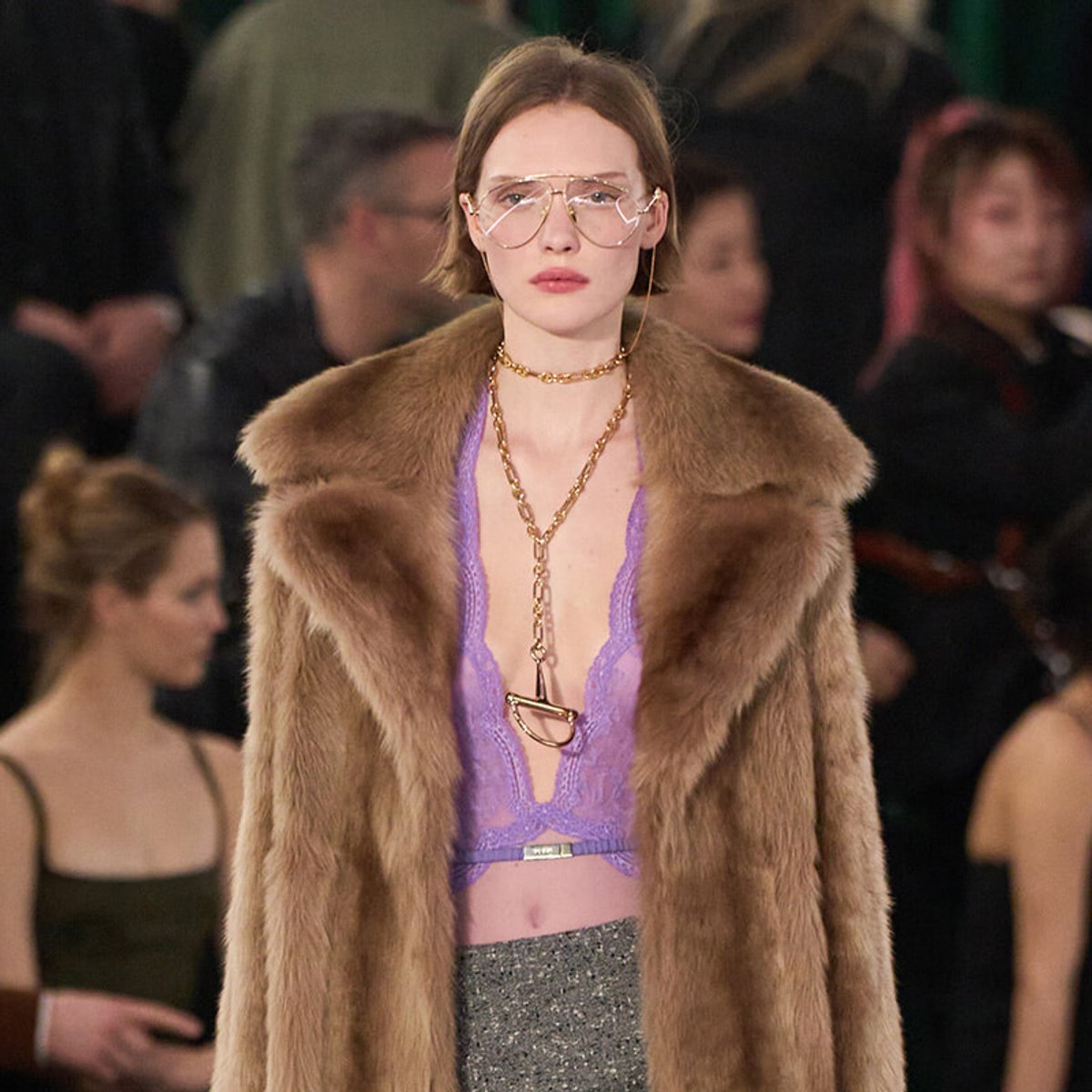 Runway model in a brown fur coat over a lilac lace bralette, with a glittery gray skirt and gold jewelry, wearing large clear glasses.