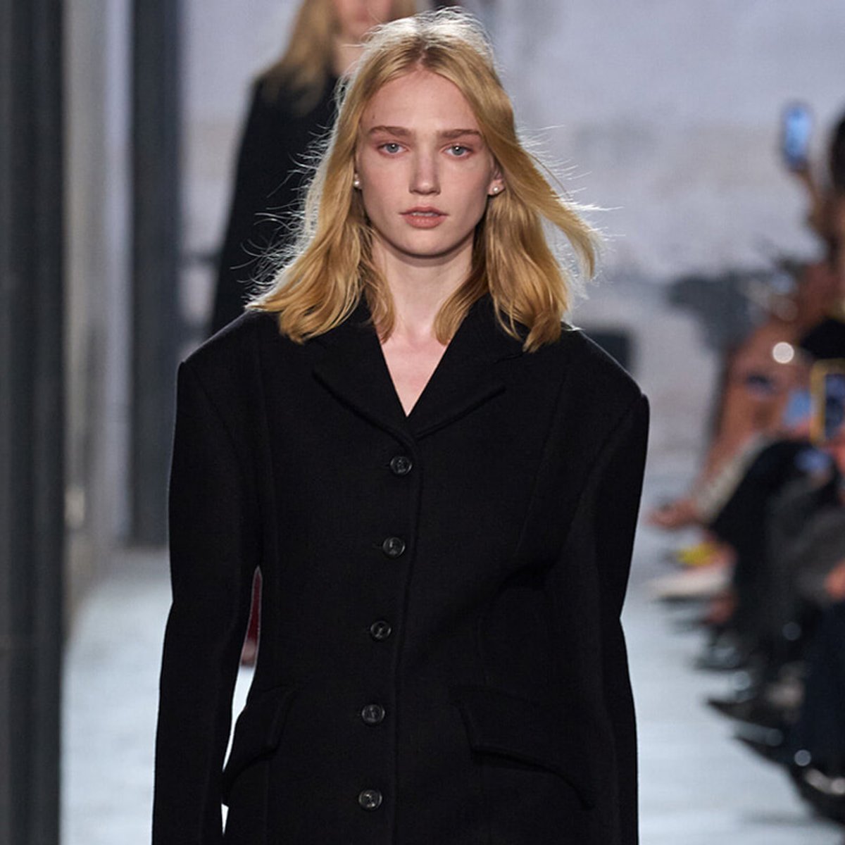 Female model walking a runway in a black tailored coat during a fashion show, neutral expression visible, blurred audience in background