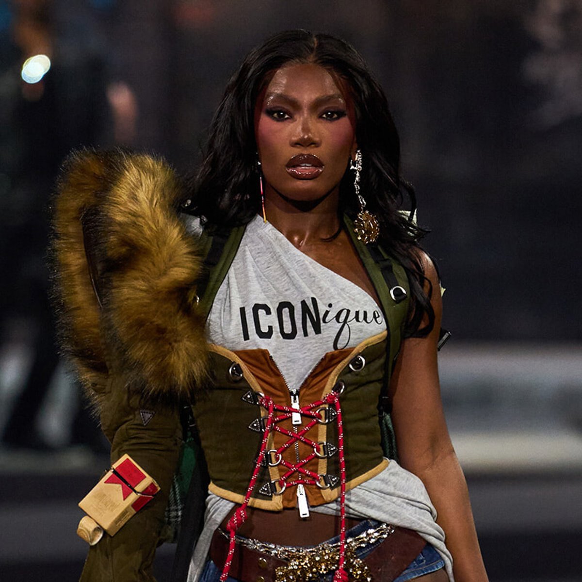 Model walking runway in edgy, mixed-media outfit with a fur-trimmed coat and corset-laced top, bold makeup and statement jewelry