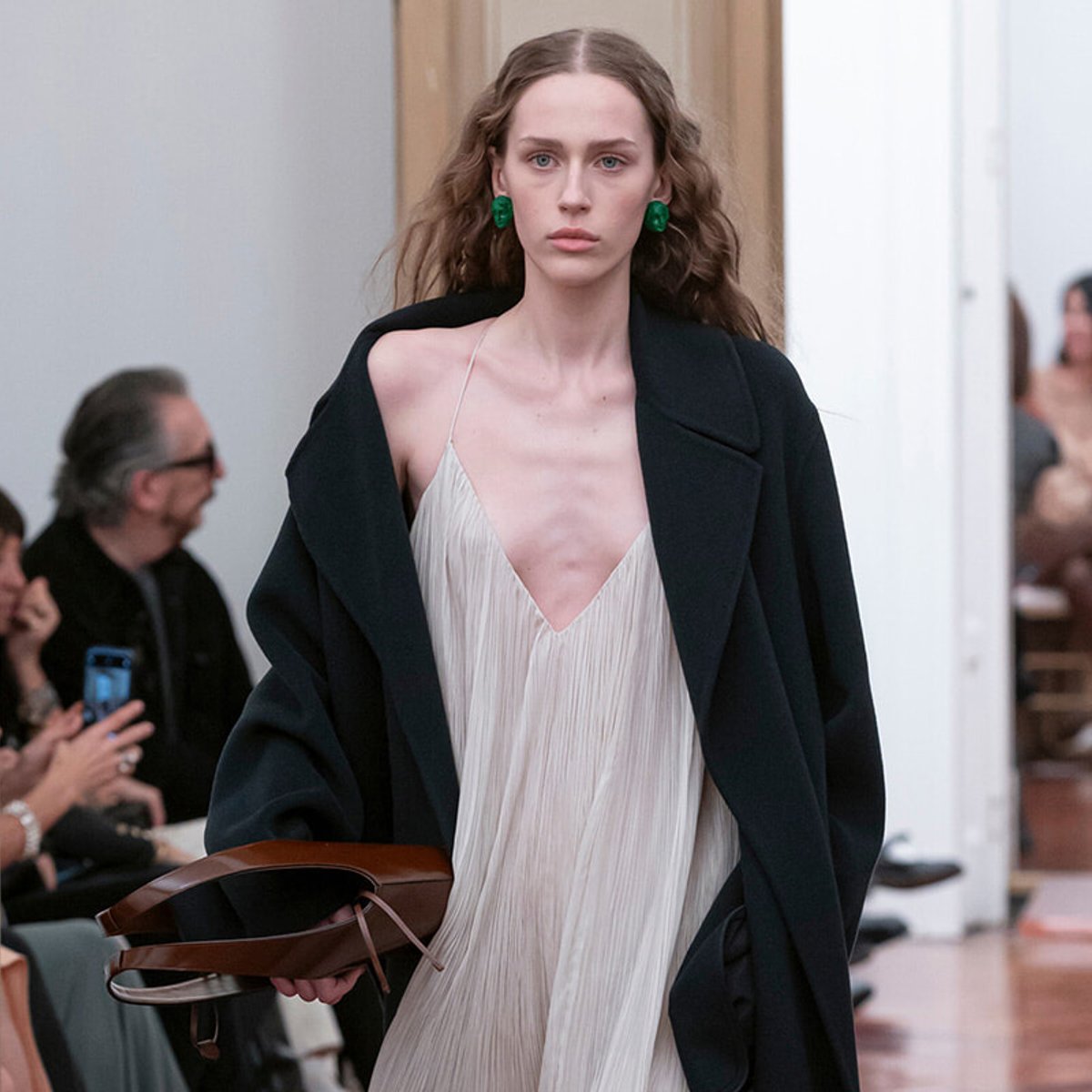 Model walking the runway in a cream pleated slip dress under a black coat, green earrings, and a brown handbag in hand.