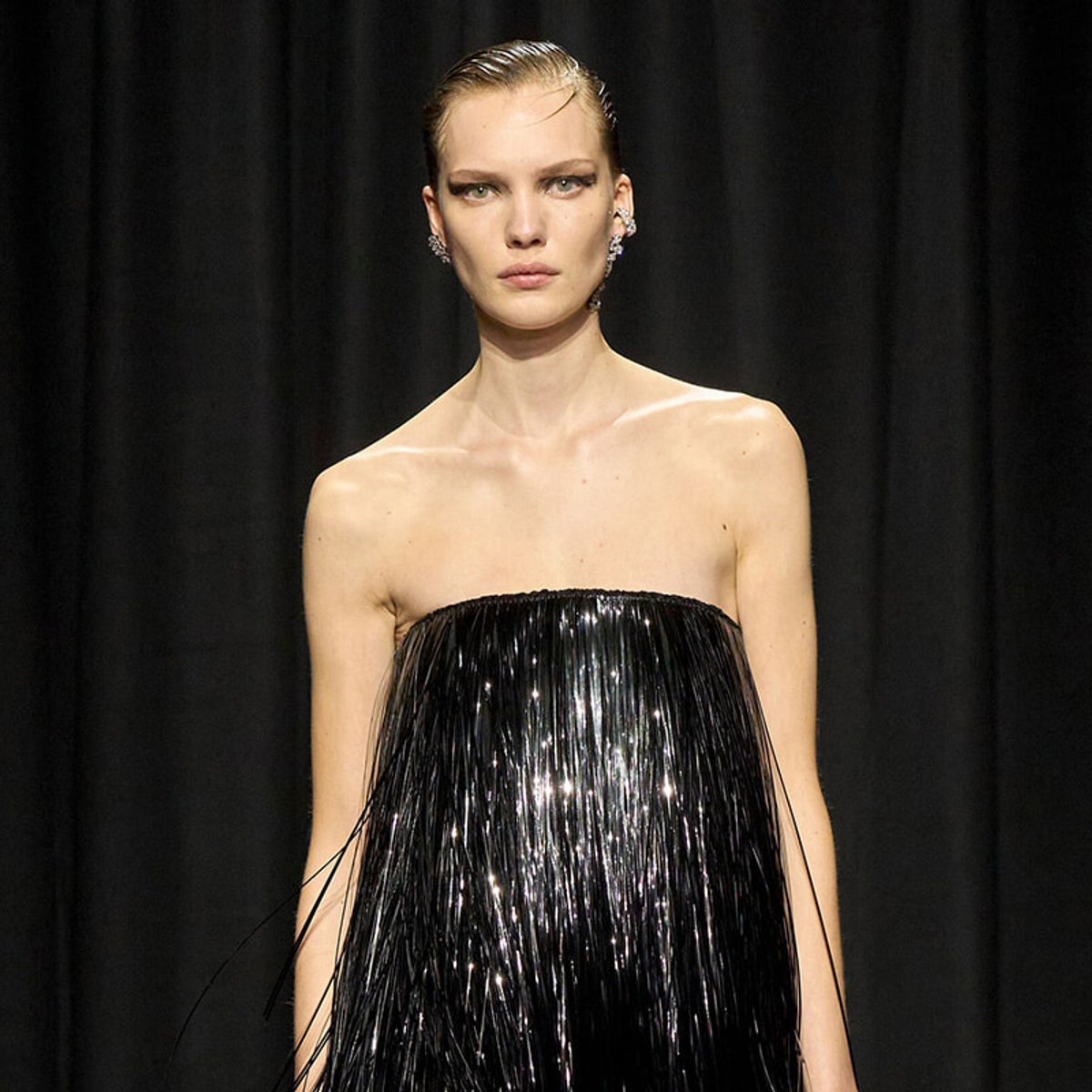 Model on a runway wearing a strapless black fringe dress against a dark backdrop.