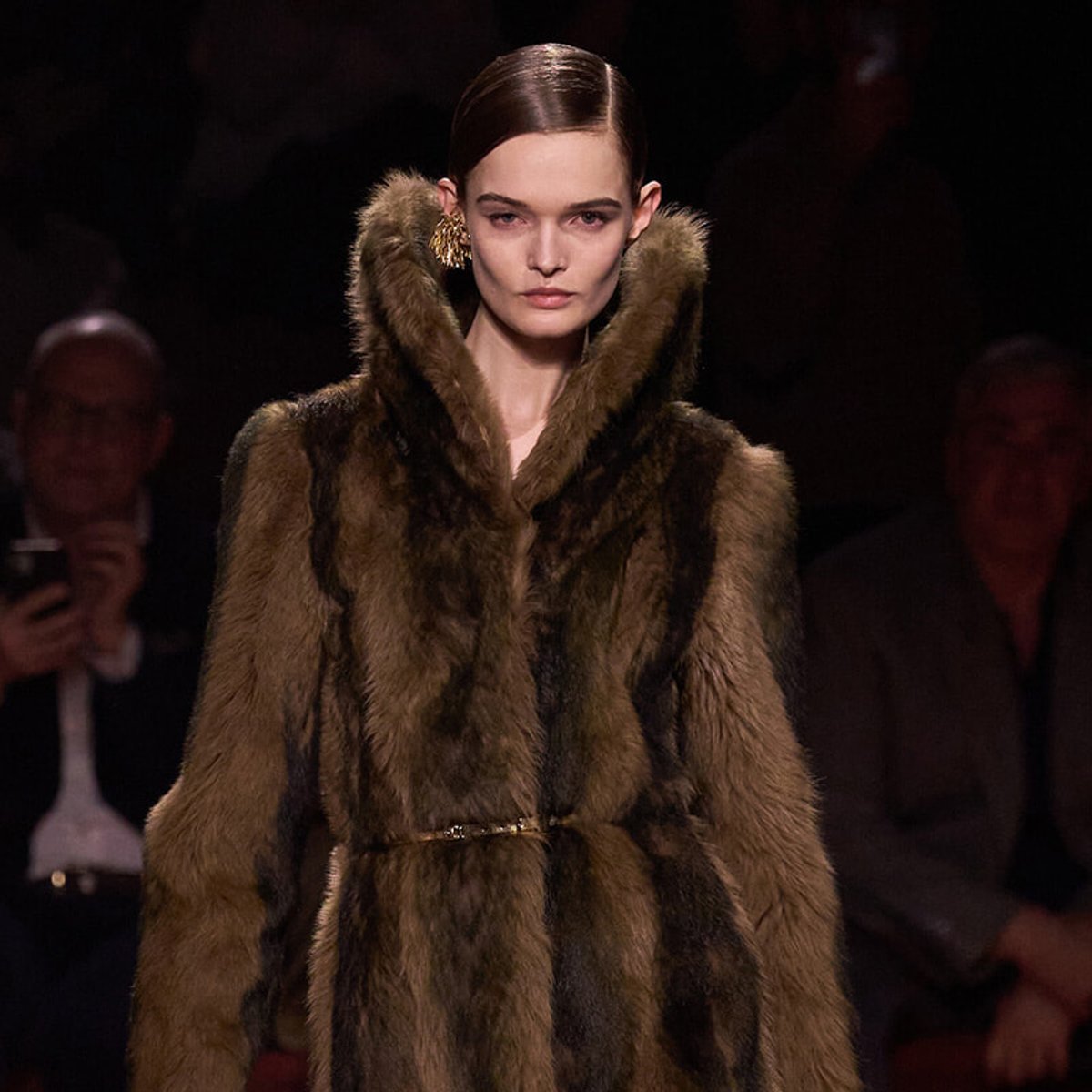 Model walking the runway in a brown fur coat with a slim belt, center-stage during a fashion show.