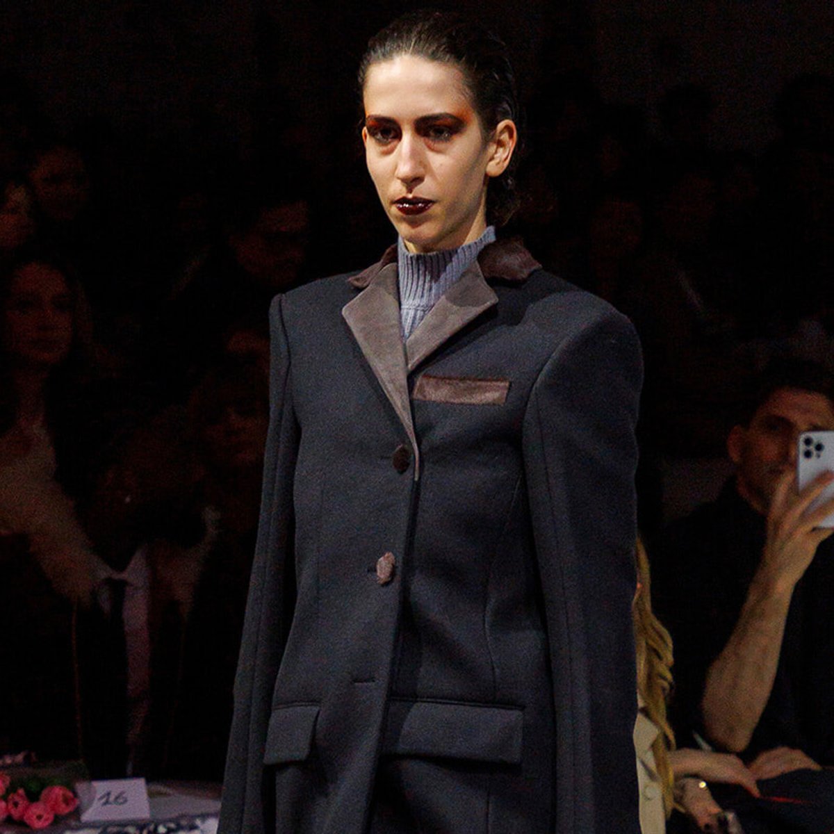Model walking the runway in a dark tailored coat with brown lapels and a light gray turtleneck underneath.