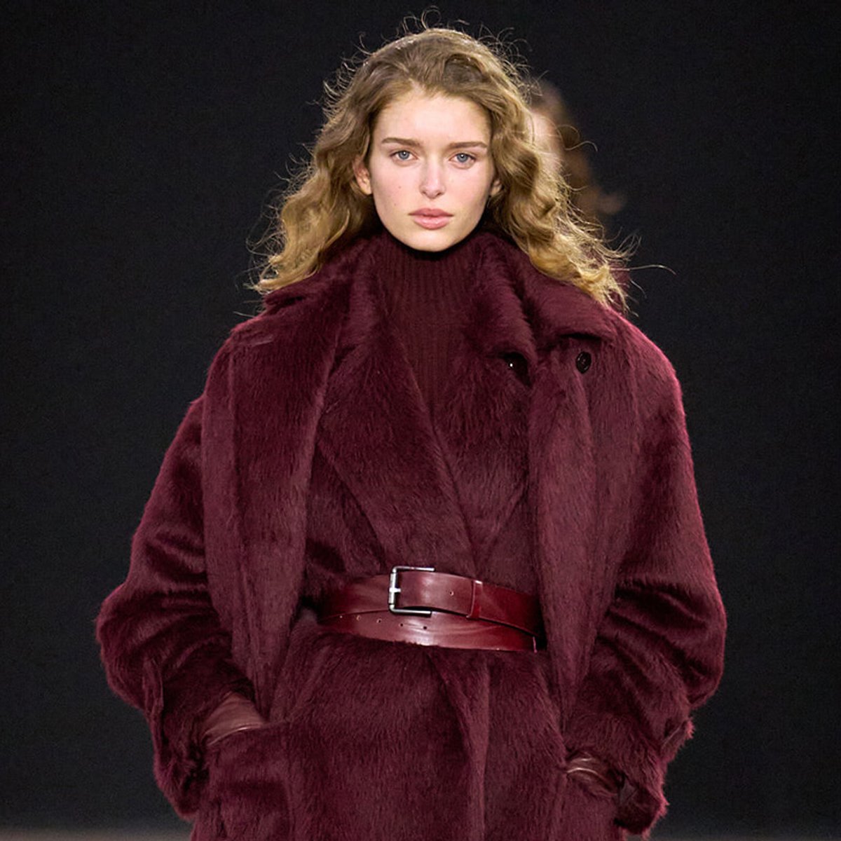Female model walking a fashion runway in a burgundy faux fur coat cinched with a wide matching belt.