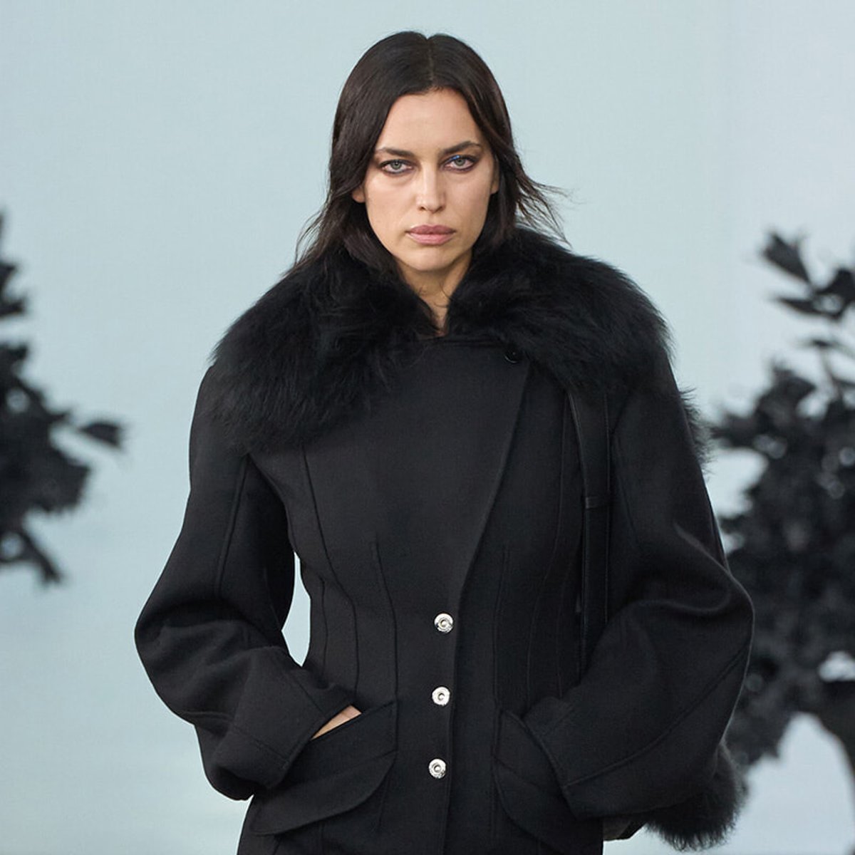 Woman modeling a black coat with a thick fur collar, hands in pockets, neutral expression.