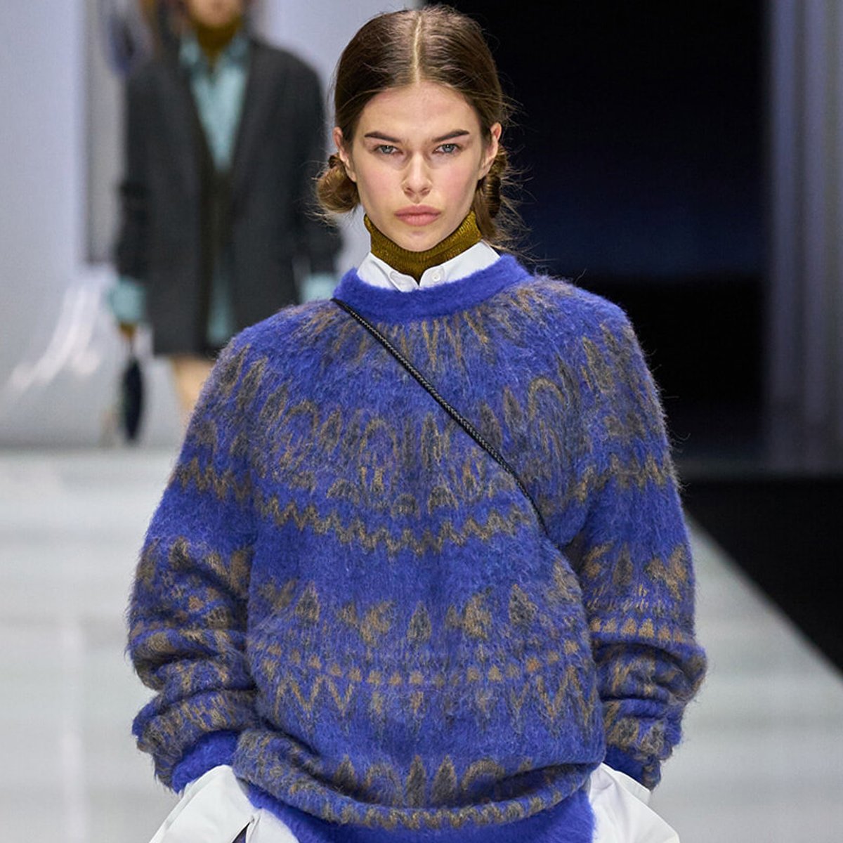 Model on a runway wearing a blue and brown fuzzy patterned sweater over a white shirt, with a thin crossbody strap visible
