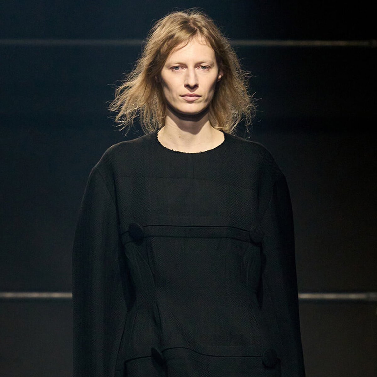 Female model on a dark runway wearing a black textured long-sleeve dress with a round neckline under spotlight.