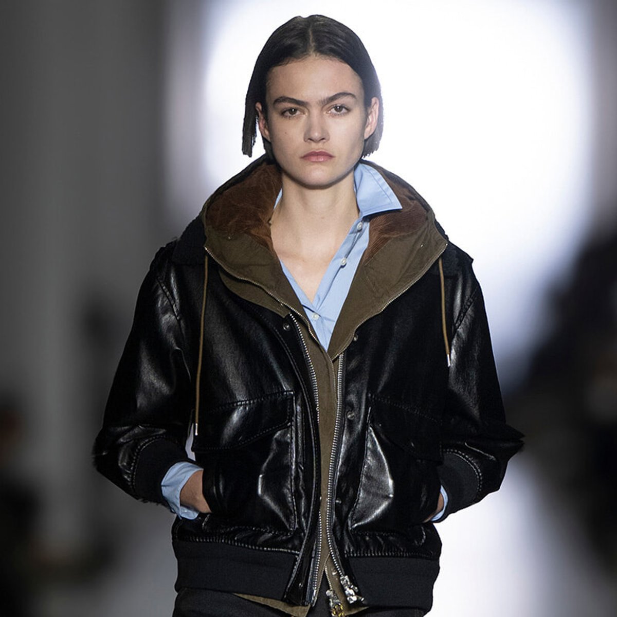 Female runway model wearing a shiny black leather bomber jacket over a light blue button-down shirt, with brown inner lining and hands in pockets.