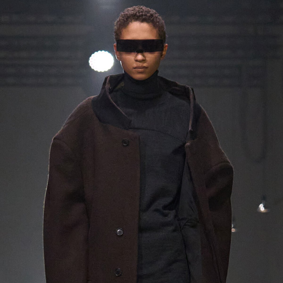 Model walks a runway in a dark turtleneck under an oversized brown coat with a black eye-covering visor across the eyes.