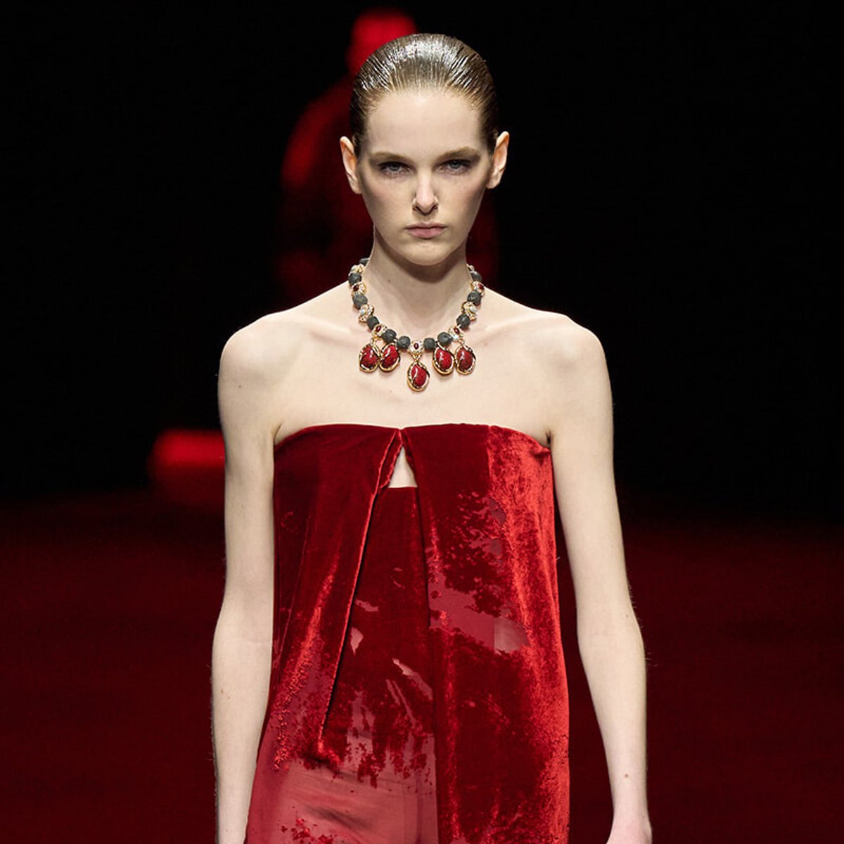 Model on a runway wearing a strapless red velvet dress with a statement necklace, a dark background behind her.