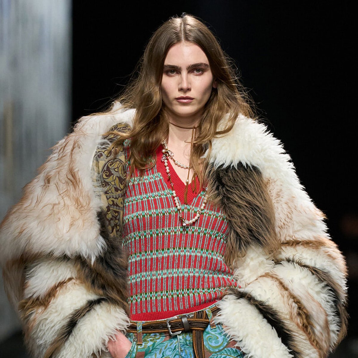 Model on the runway wearing a voluminous fur coat over a red plaid top and turquoise patterned pants, with layered necklaces.