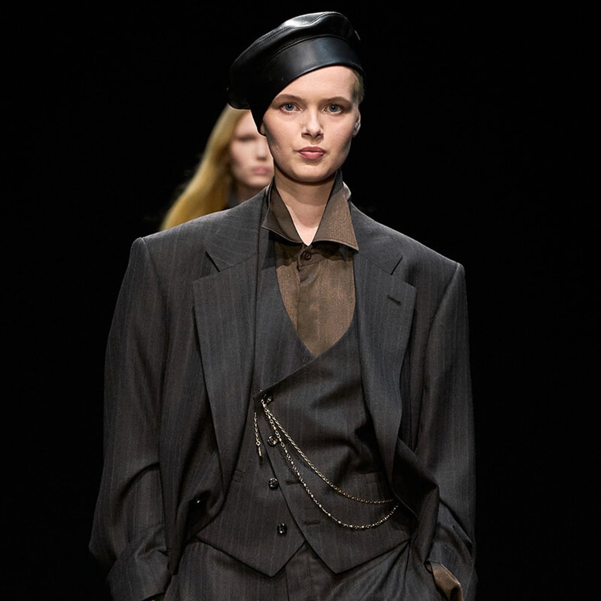 Model on a fashion runway wearing a dark pinstripe suit, brown shirt, and a black leather beret with a chain accessory.