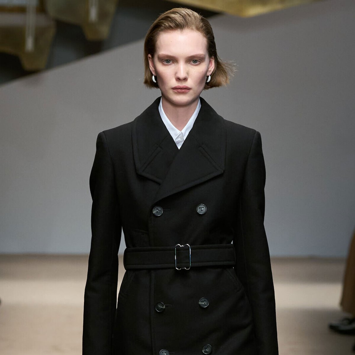 Model wearing a black double-breasted belted coat with a white collared shirt, short blonde hair, and a neutral expression on a runway.