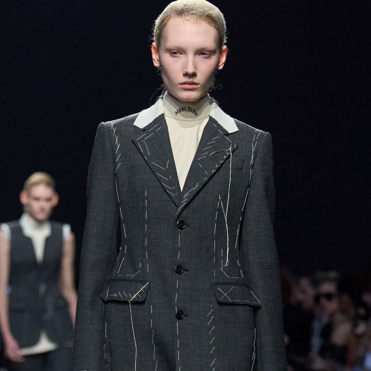 Model walking a fashion show in a dark gray blazer with white collar, white stitching details, and a cream turtleneck with Moschino logo visible at the neck.