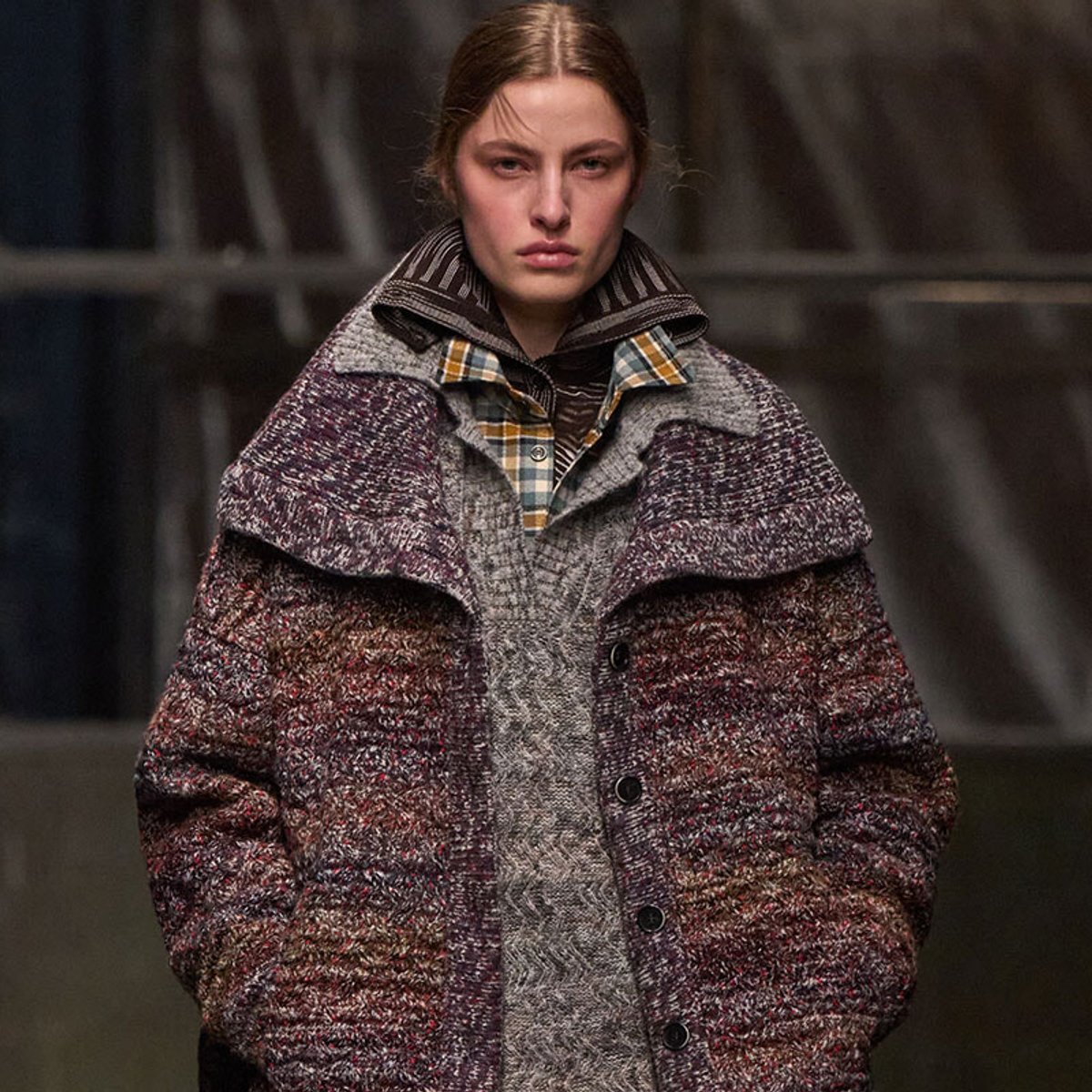 Model wearing a thick, multi-textured wool coat with a plaid shirt peeking from underneath, facing the camera.