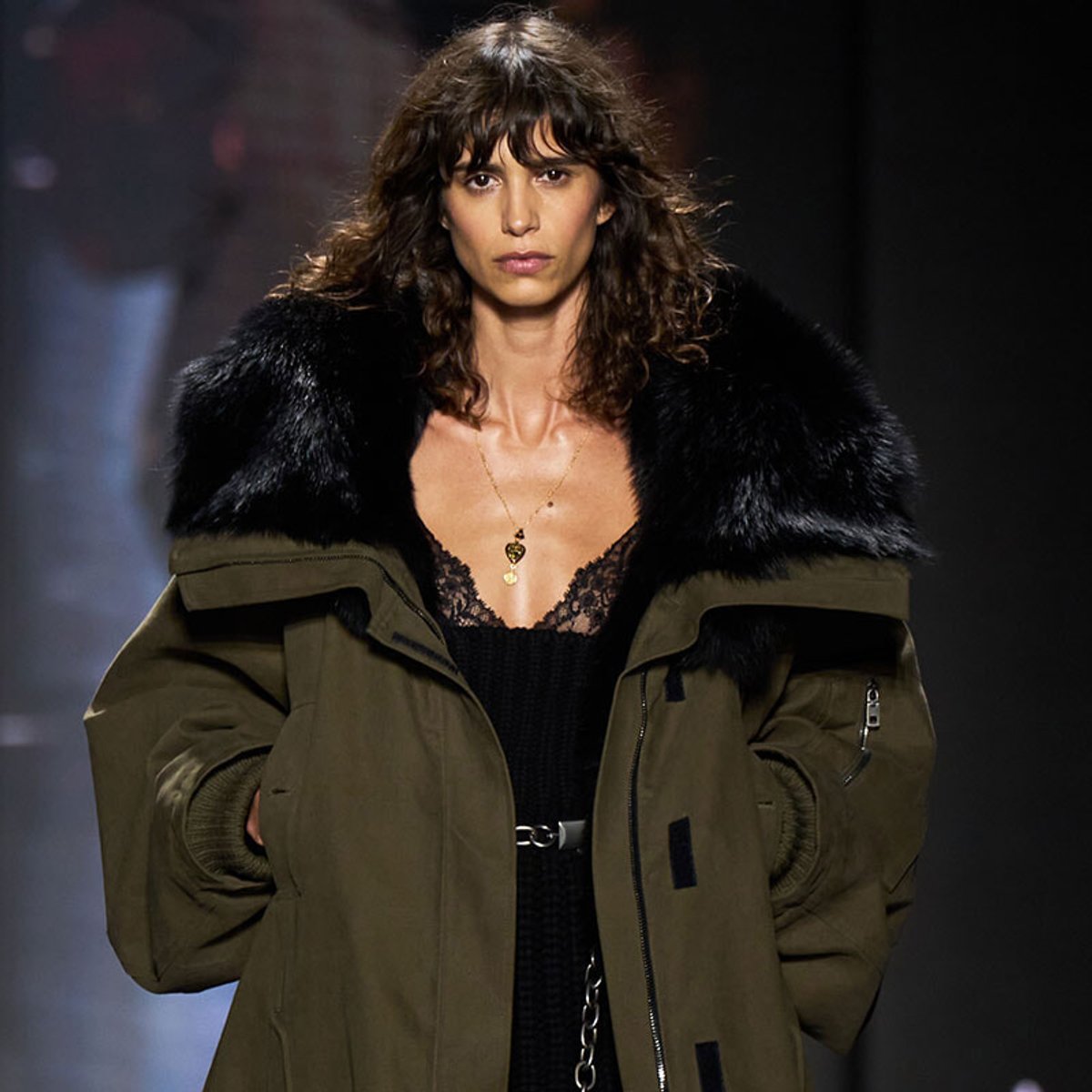 Model walking the runway in an oversized olive parka with a black fur collar, wearing a black lace top and a pendant necklace.