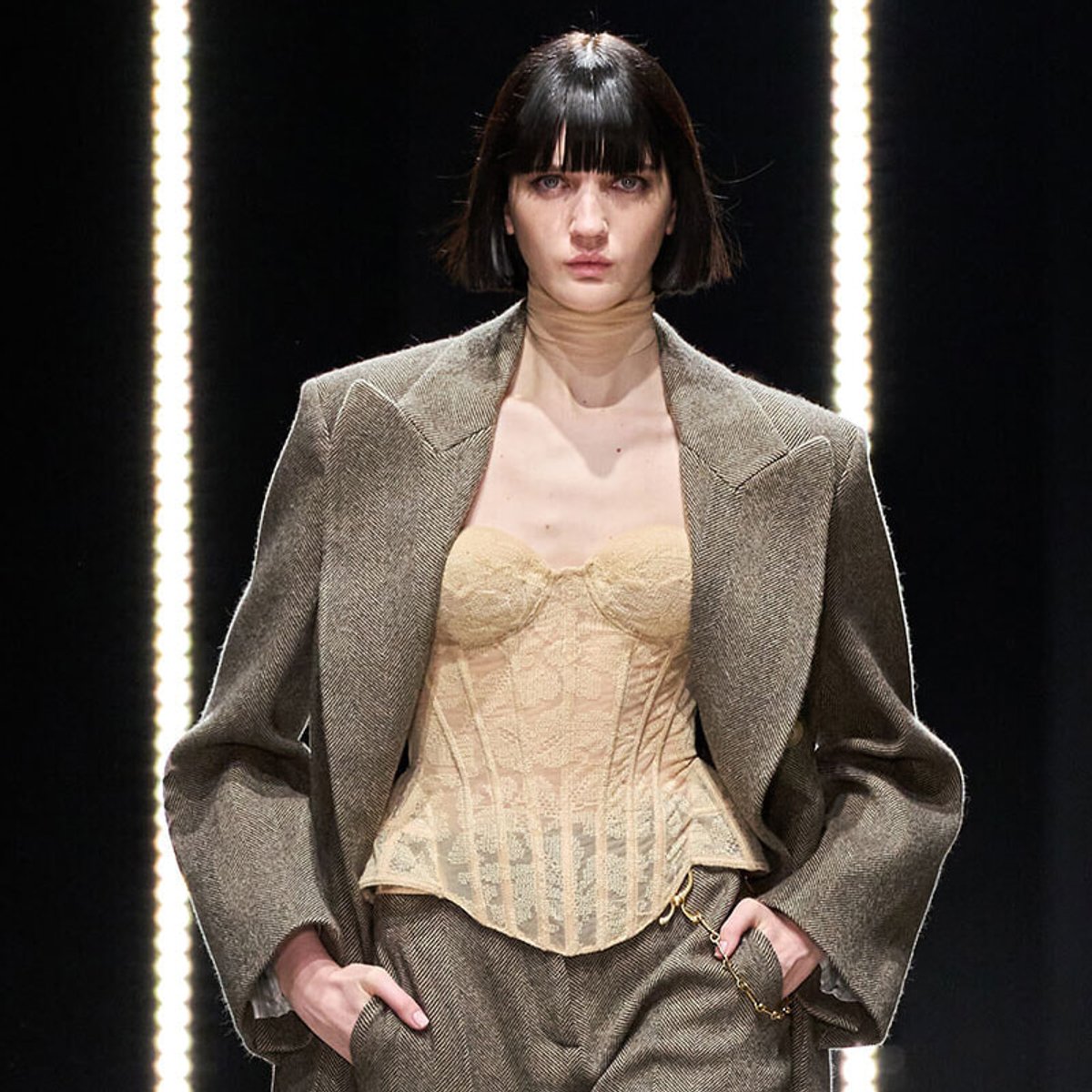 Model on runway wearing an oversized brown tweed blazer over a beige corset, with hands in pockets and bright lights in the background.