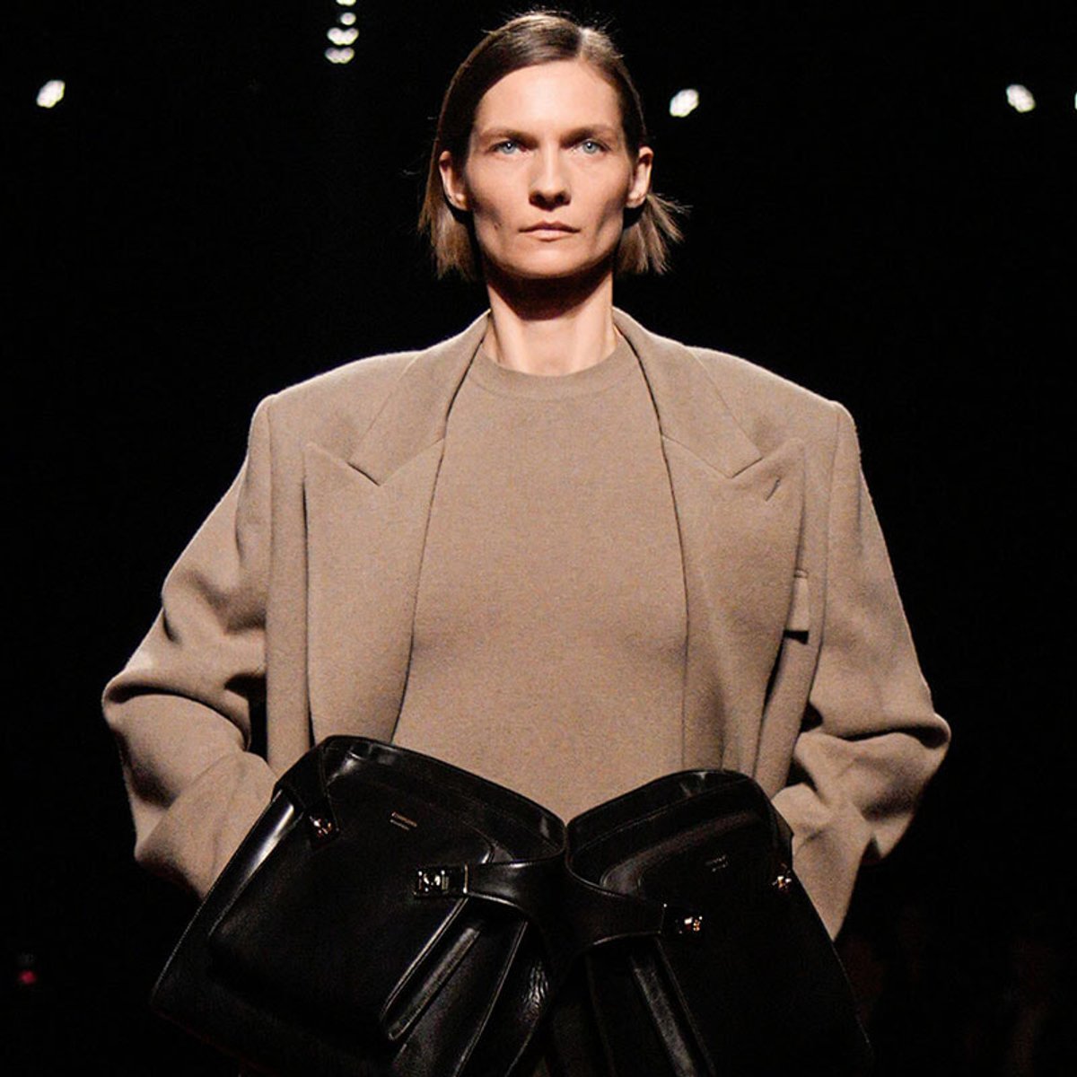 Model on a fashion runway wearing a loose beige coat over a matching top, holding two large black leather bags at the front