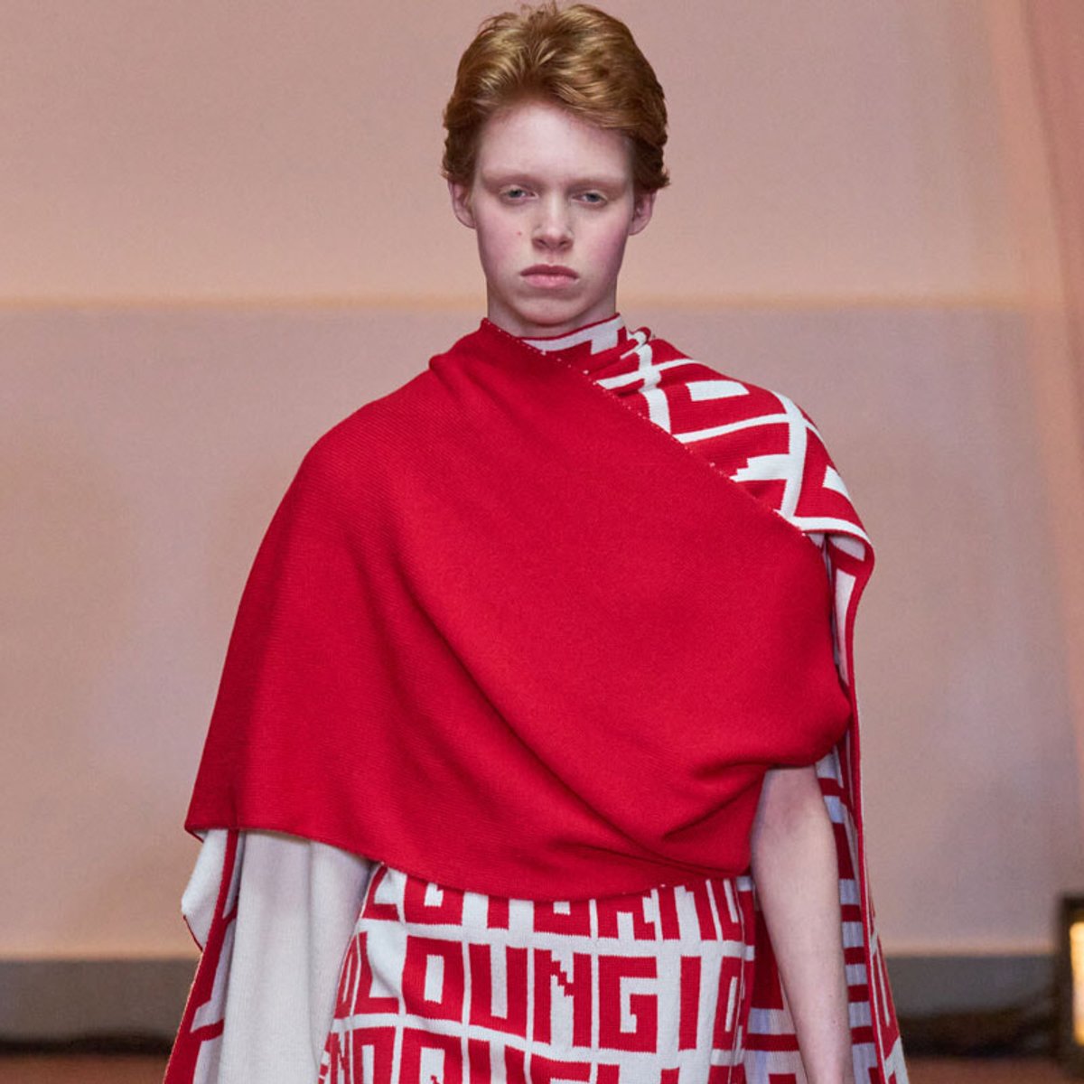 Runway model wearing a bright red poncho over a white and red geometric-pattern dress.