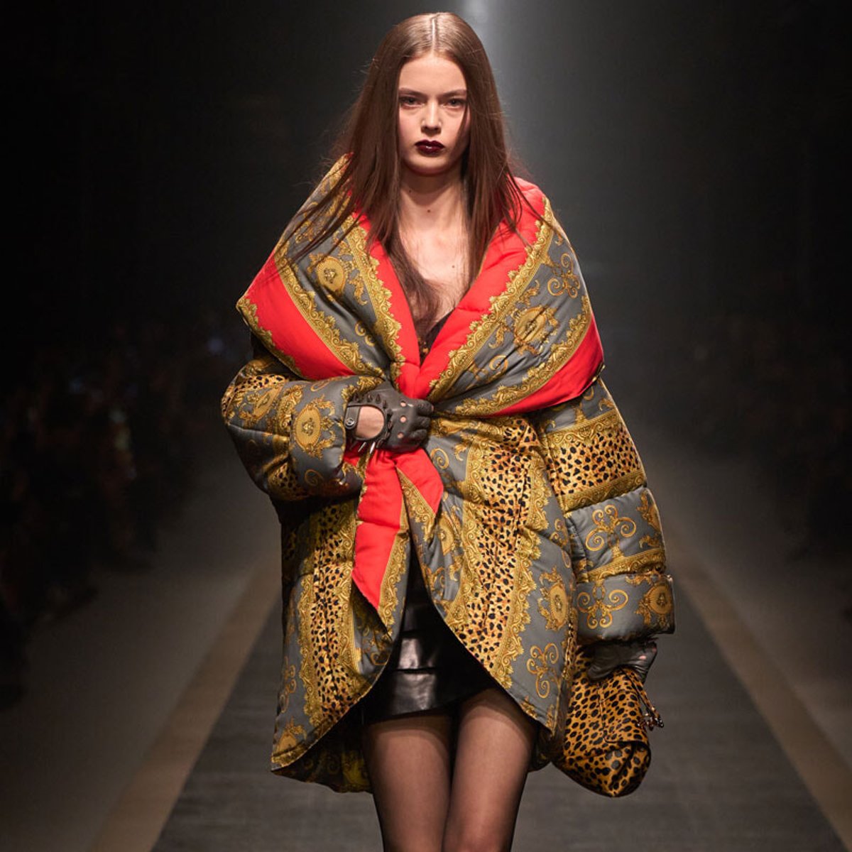 Model walking a runway in a large, ornate quilted coat with red and gold baroque patterns.