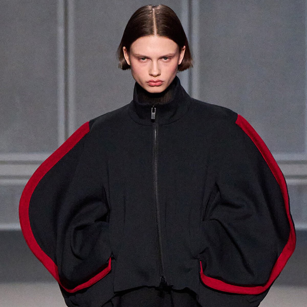 Female model walks runway in an oversized black jacket with red piping along the sleeves.