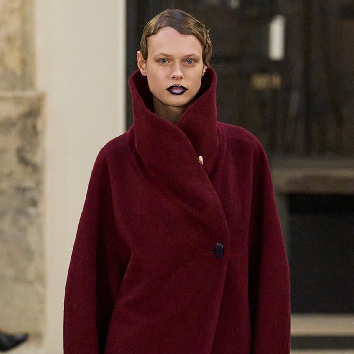 Model walking in a burgundy oversized coat with a high, structured collar and a single button closure at the chest.