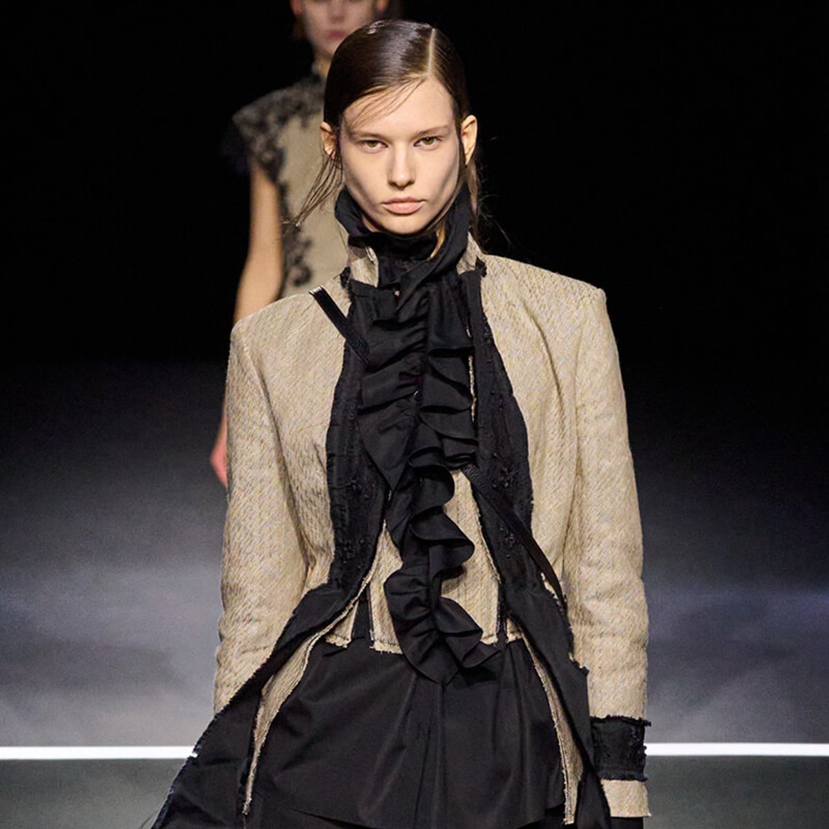 Model walking the runway in a beige textured blazer over a black ruffled blouse and skirt, with a dark backdrop behind.