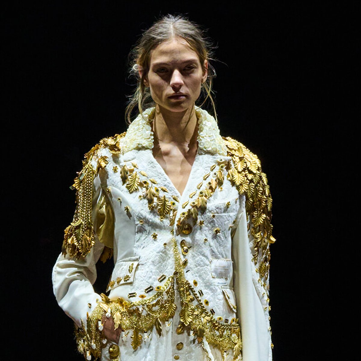 Model on a dark stage wearing a white suit adorned with gold leaf embroidery and epaulettes.