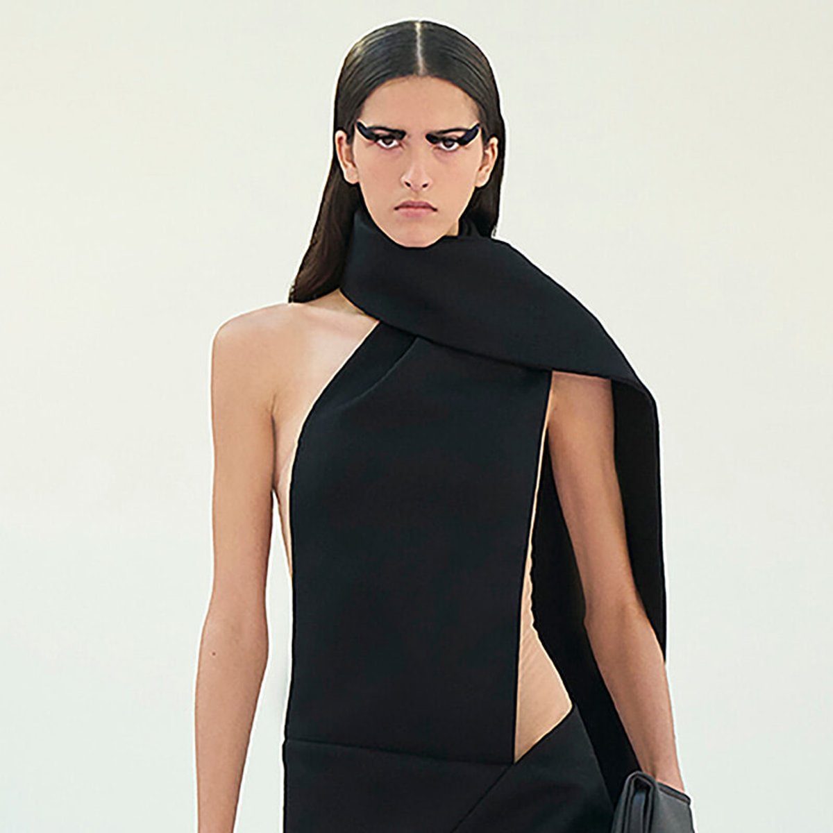 Model wearing a black asymmetrical dress with a wide scarf-like collar against a pale background, bold eye makeup emphasized.