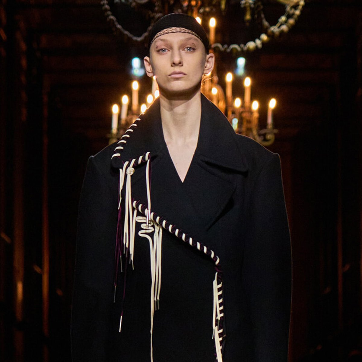 Model wearing a black coat with white braided cords on a dark runway, candlelit backdrop behind.