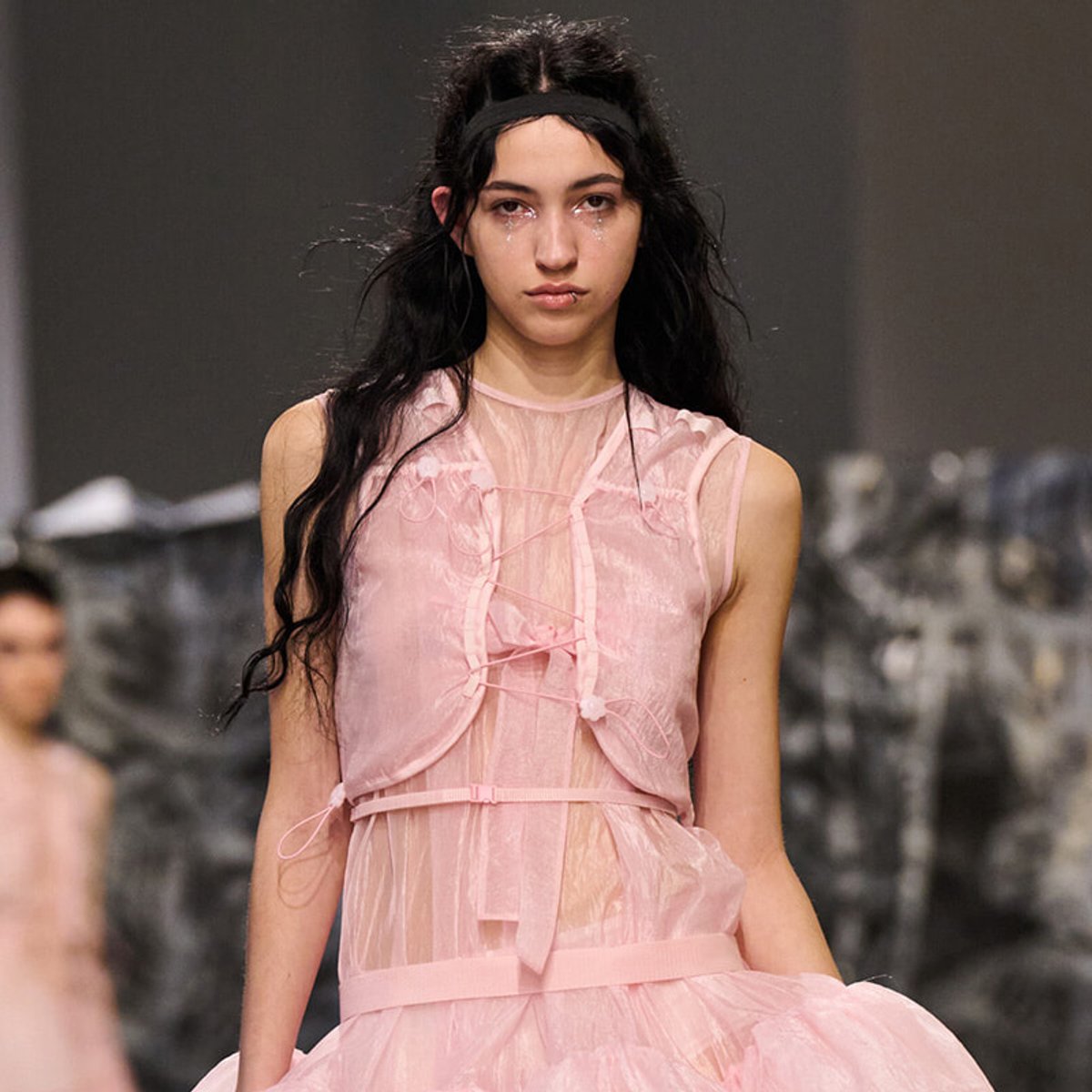 Model walking the runway in a pink sheer dress with lace details and a black headband to hold back her hair.