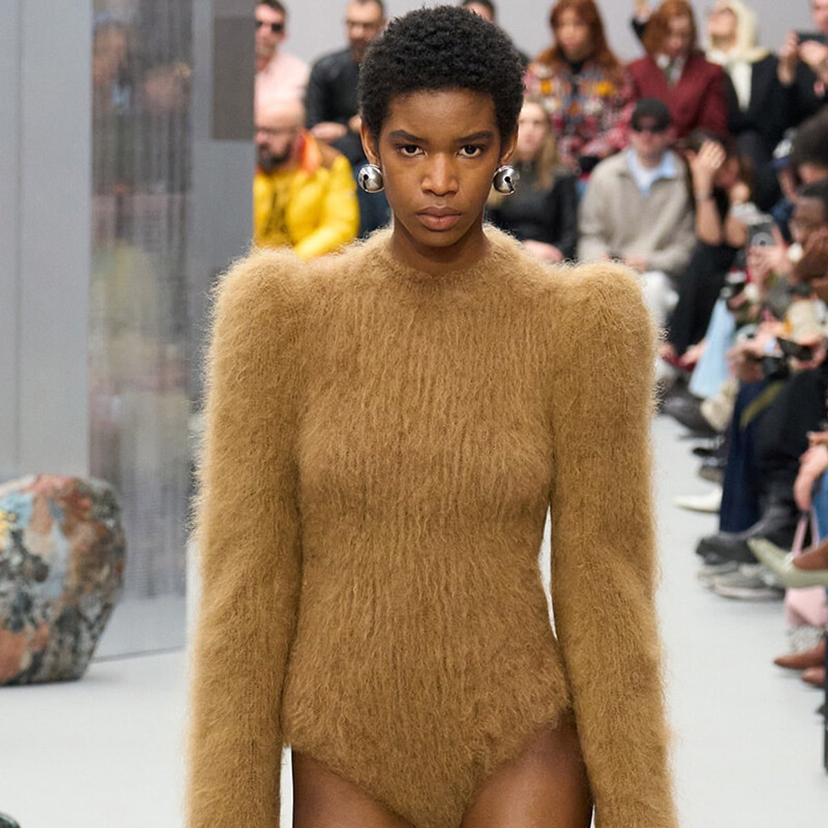 Model on a runway wearing a camel-colored fuzzy knit bodysuit, audience blurred in the background.