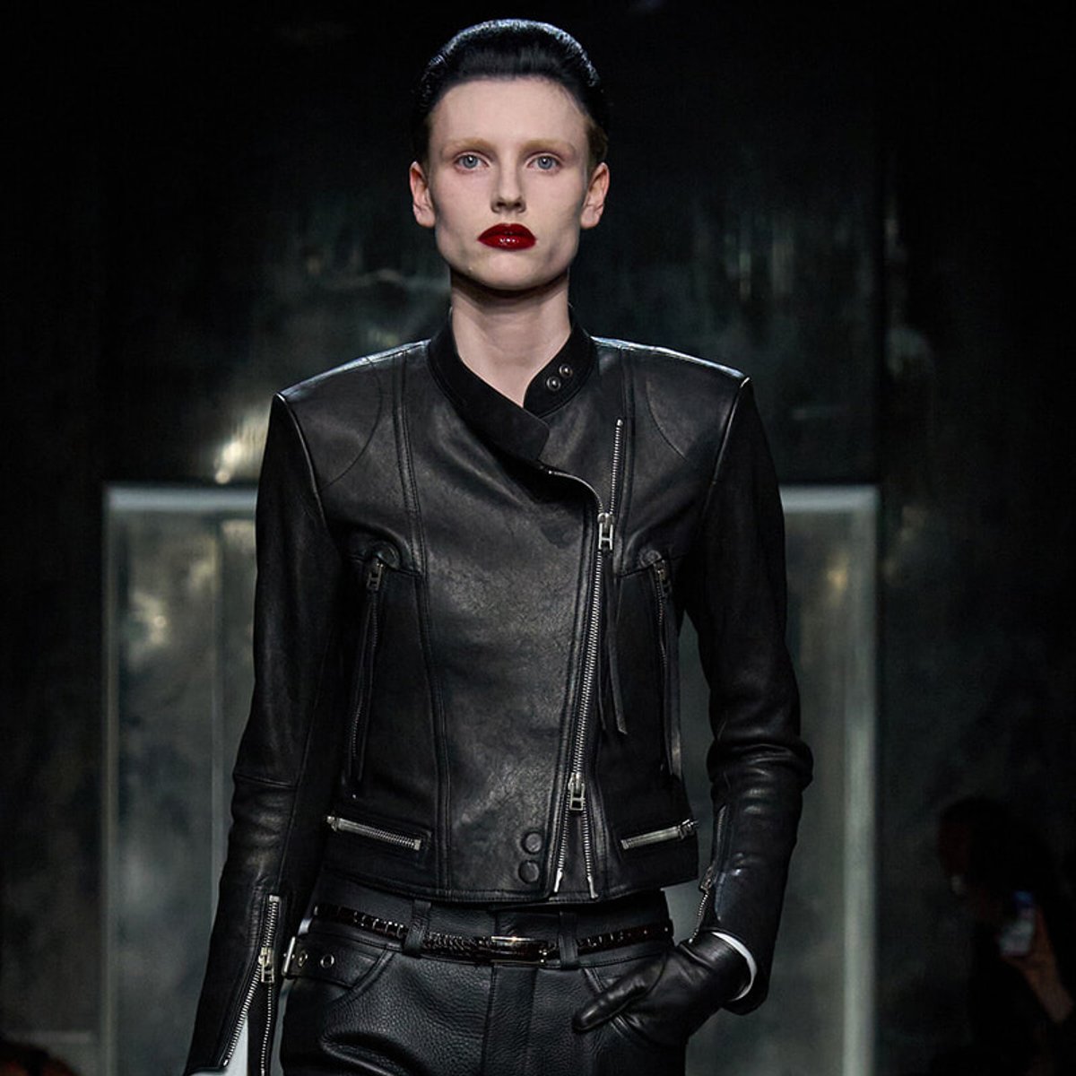 Model wearing a black leather biker jacket on a runway, hands in pockets, bold red lipstick, dark backdrop behind.