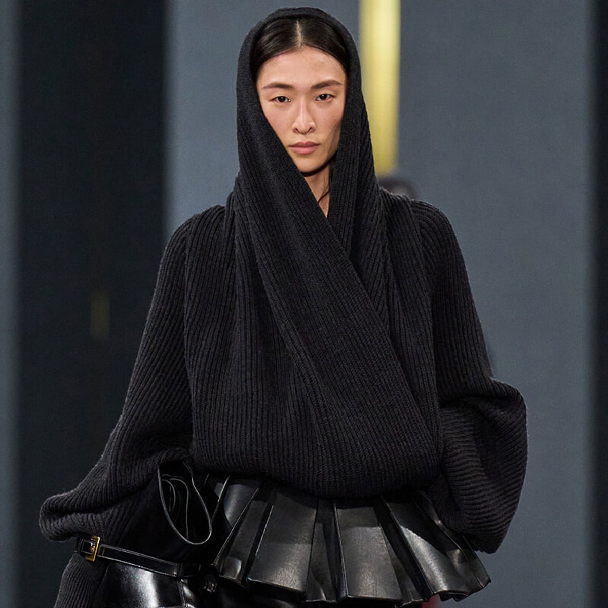 Model walking the runway in a black oversized ribbed knit hoodie and black belted skirt, minimalist futuristic look.