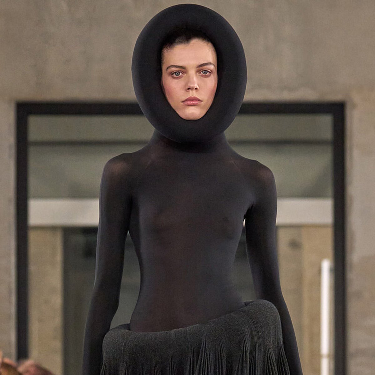Model on a runway wearing a black full-body suit with a large circular hood and fringe skirt at a fashion show.