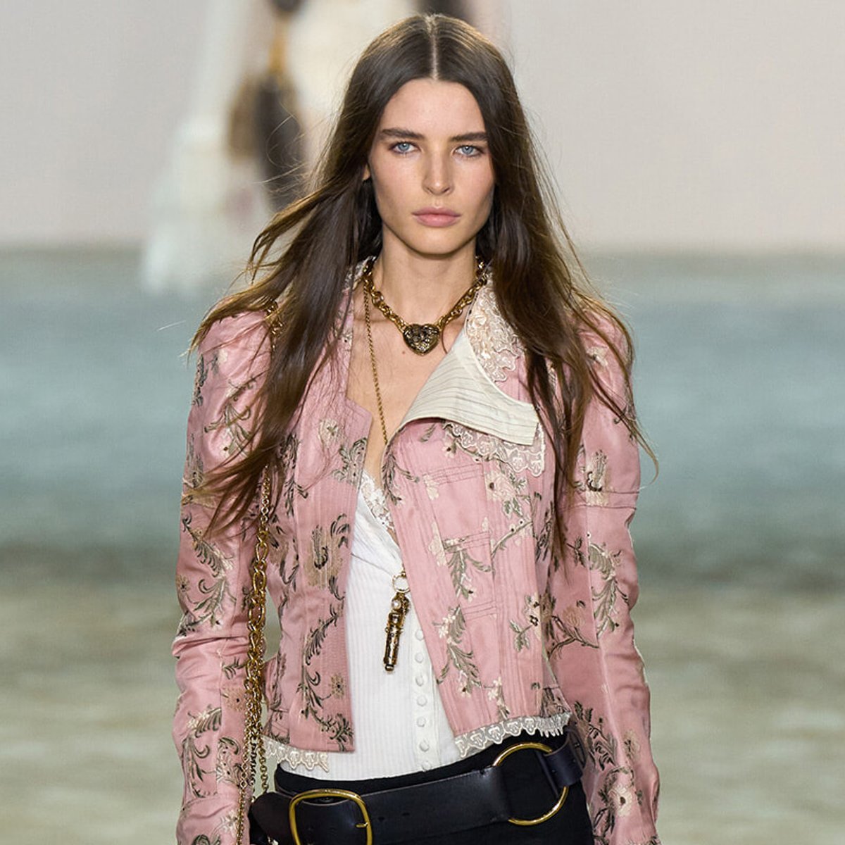 Model walking runway in a pink floral embroidered jacket, white top, and chunky belt with gold accents.