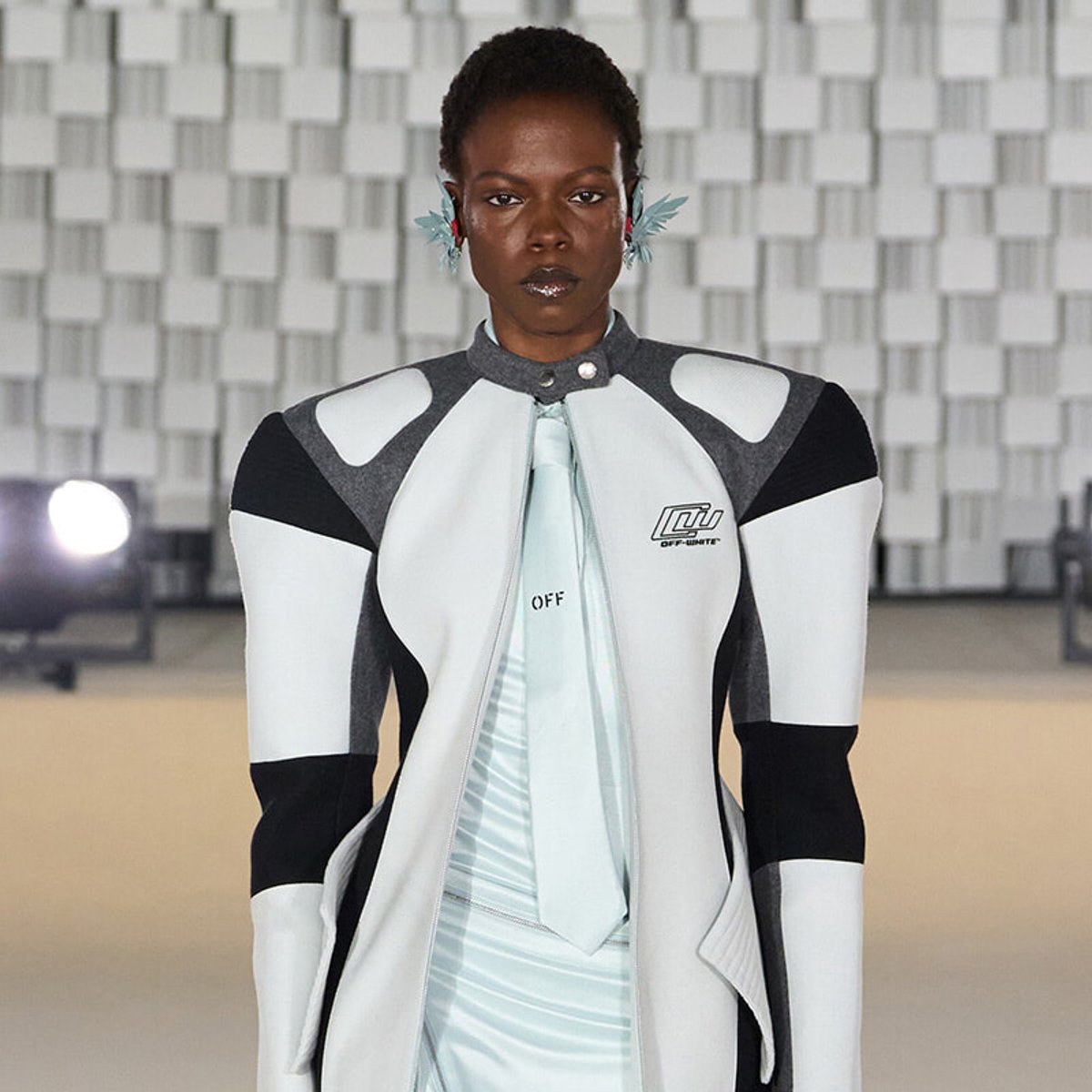 Female model on a runway wearing a white and gray geometric jacket with a mint tie and visible Off-White logo.